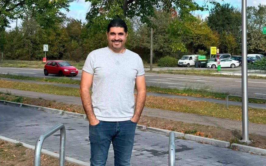 Husam Noaman standing and smiling on a sunny street, with a large tree behind him