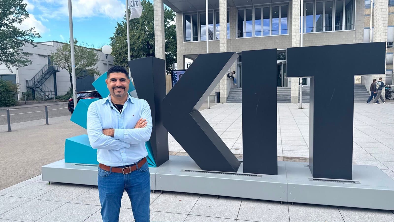 Husam Noaman standing with his arms crossed in front of the large KIT sign at the Karlsruhe Institute of Technology